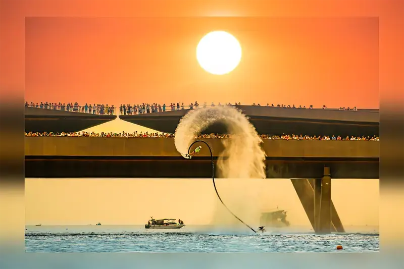 Water-powered performer in front of Kiss Bridge beneath an orange sky, with spectators watching from the bridge