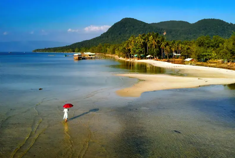 Wide view of Rach Vem coast, sandbars, shoreline structures and a person carrying a red umbrella