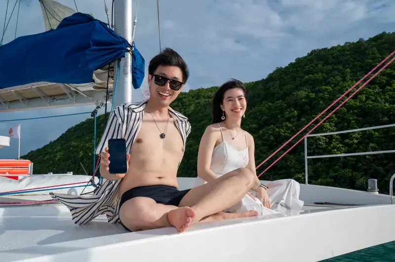 Cặp đôi tươi cười ngồi cạnh nhau trên thuyền buồm, phía sau là bờ biển xanh cây.