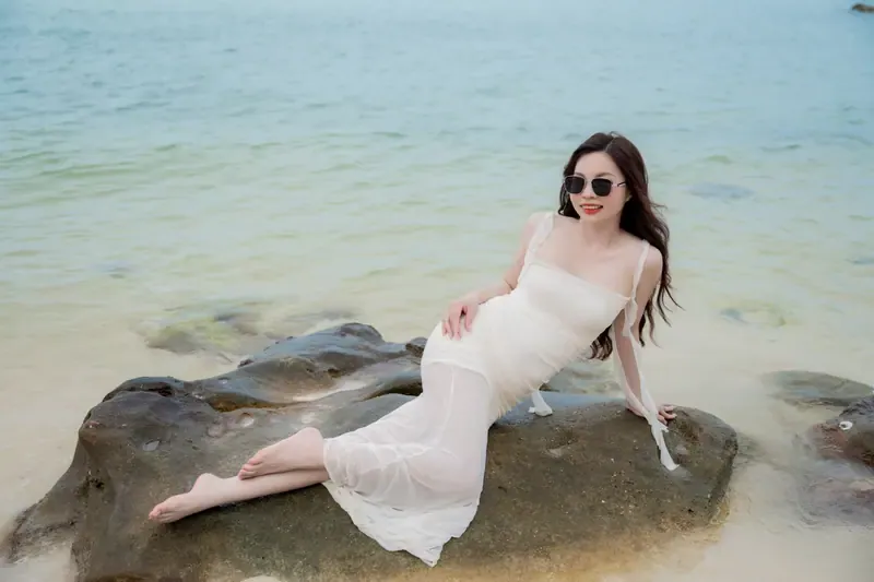 A woman in a white dress and sunglasses reclining on a shoreline rock beside pale turquoise water.