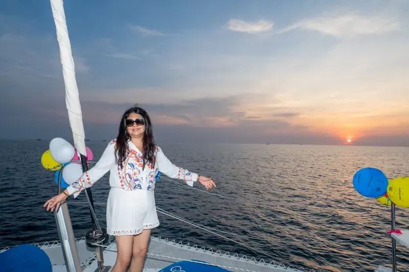 A woman in sunglasses and a white embroidered outfit standing on a boat deck with balloons, the sea and the setting sun behind her.