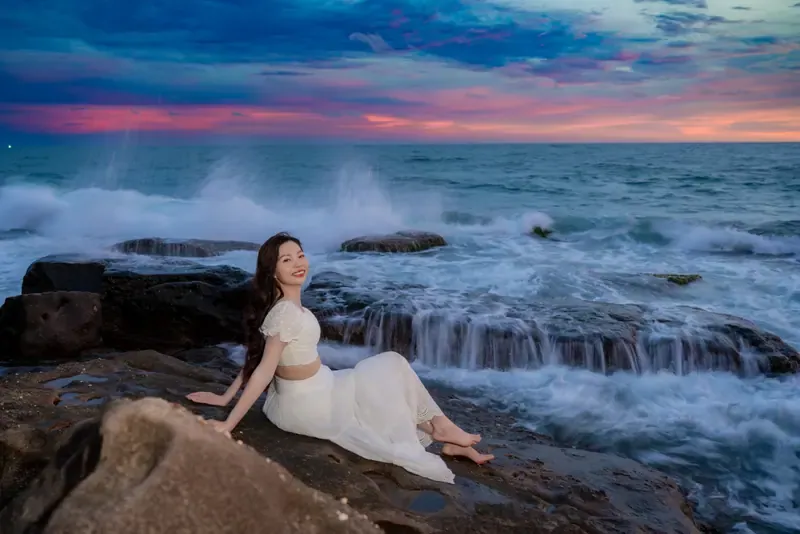 A woman in white sitting on coastal rocks with breaking waves and a pink and blue evening sky behind her.