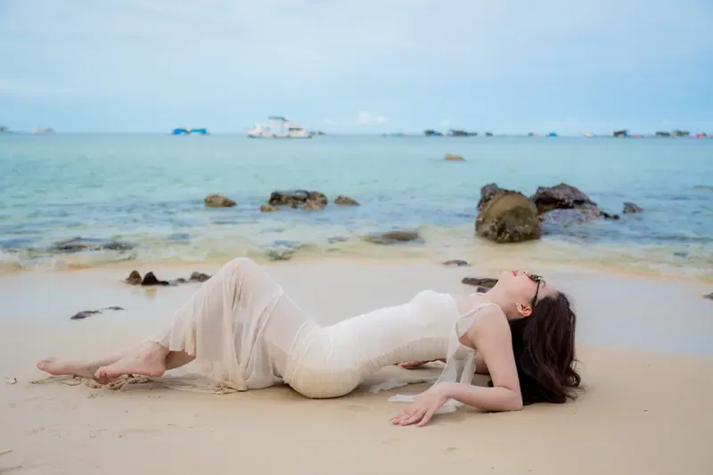 Một người phụ nữ mặc váy trắng nằm trên cát, bên làn nước xanh và những tảng đá ven bờ.