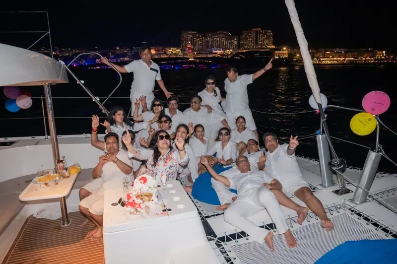 Guests gathered around a cake on a boat at night, with balloons and an illuminated waterfront behind them.
