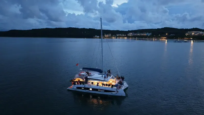 A wide aerial view of a catamaran with warm cabin lights in a bay at dusk, with hills and clouds in the distance.