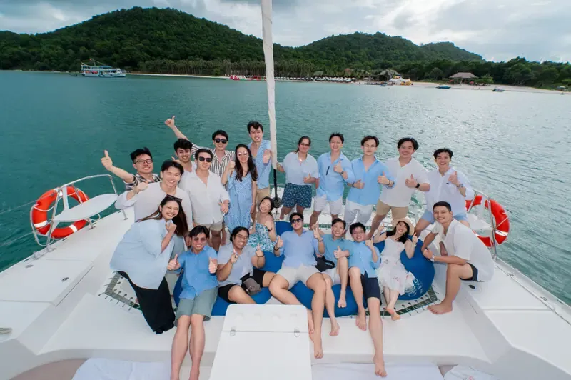 A group in blue and white smiling together on a catamaran, with a green shoreline behind them.
