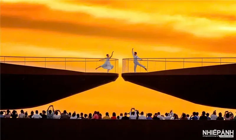Hai người tạo dáng ở hai đầu Cầu Hôn dưới bầu trời cam, phía dưới có khán giả.