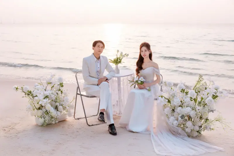 Bride and groom seated at a lace-covered table between white floral arrangements on the beach.