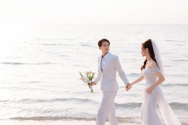 Couple in white wedding attire holding hands as they walk beside the waves.