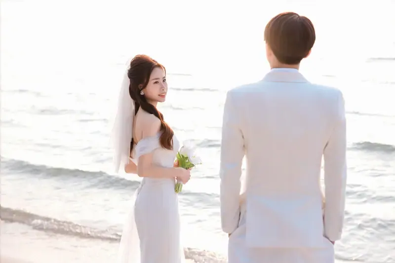 Bride holding pale tulips looking towards the groom beside the sea.